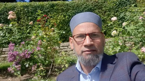 Muhammed Rashid Muhammed Rashid takes a selfie in a garden, he is wearing a blue plaid jacket, blue shirt and kufi