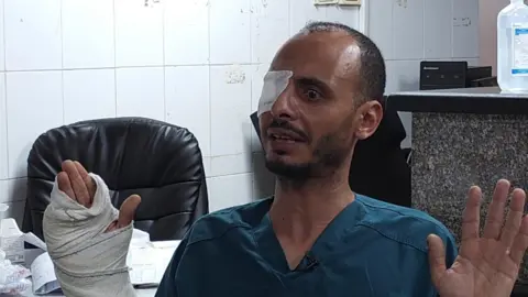 Ashraf al-Attar sat in a turquoise hospital shirt, his hands raised in the air in front of him. His right hand was wrapped in heavy bandages, his fingers and thumb protruding. His right eye was covered with a patch.