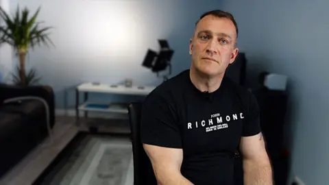 A well-built man in his late 30s sitting in a chair wearing a black t-shirt and looking at the camera. He is sitting in a grey-painted room with a back-lit wall and a plant in the corner. He is not smiling.