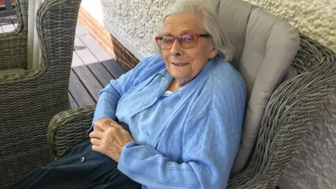 Elderly Valerie Knights sits in a garden chair and looks at the camera. She is smiling.