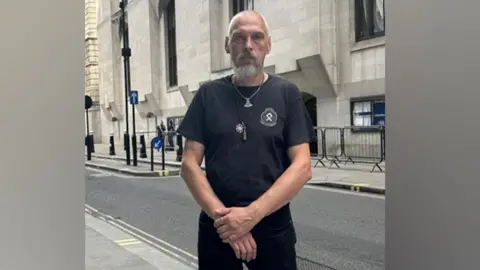 CPS Piotr Kucharski standing outside the Central Criminal Court. He is wearing a black T-shirt and trousers and has a couple of necklaces. He is tanned with a shaved head and grey beard and moustache. 