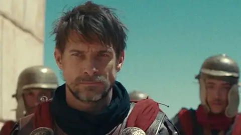 Netflix A man in Roman Military armour, with a beard and red fabric over his breastplate. Behind him two foot soldiers with metal helmets on.