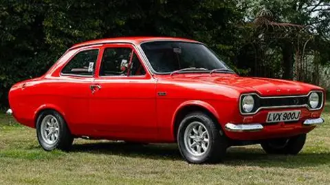 The Ford Escort Mexico, which is red, has two doors and silver lights. The picture has been taken side-on.