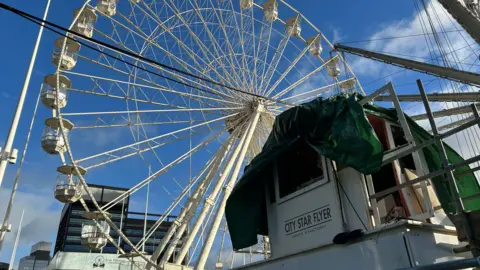 The site of the City Star Flyer after the crash. The ride can be seen cordoned off by a green fence with a big wheel in the background.