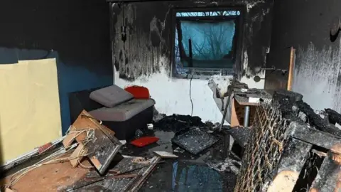 The picture show an hotel room heavily charred, with extensive fire and smoke damage covering the walls, ceiling, and furniture. Soot is thick across most surfaces. A burnt bed frame is visible on the right. The floor is strewn with debris: burnt wood, melted materials, insulation, and broken items.
