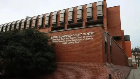 The outside of Leeds Combined Court, with white letters painted on to red bricks. There is a large green bush next to the building as well. 