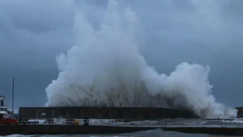 A large wave crashes against a harbour wall at Stonehaven. It is a dark, wet afternoon.