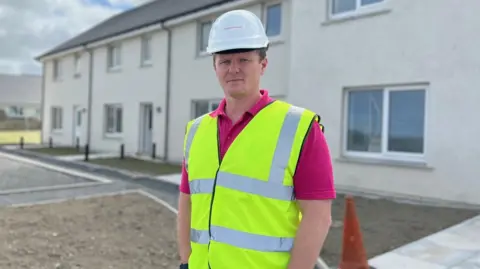 BBC Steven Kemp is standing outside a housing development. He is wearing white hard hat and a high visibility vest over a pink polo shirt.
