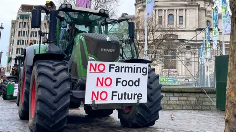 A green tractor is parked on a Westminster pavement under a grey winter sky next to a bare tree and in front of the QEII Centre. On the front of the tractor is a protest placard that reads No Farming, No Food, No Future. Pedestrians pass by.