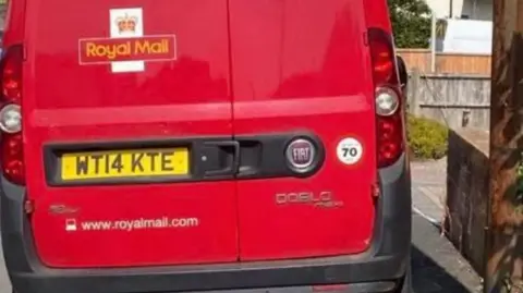 Fern Colebrook A red coloured Royal Mail van parked on a pavement right up against a brick wall.