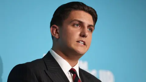 George Finch speaking at a media conference stood at a podium, wearing a black pinstriped suit