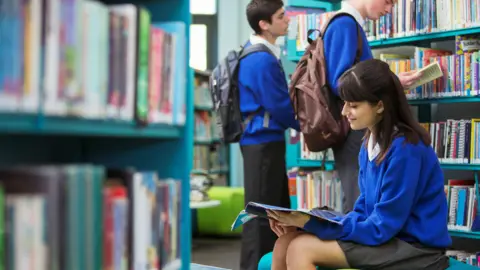 A group of pupils, wearing blue and grey school uniforms, sit in a library reading books or stand looking though titles on the shelves