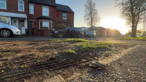 A muddy grass verge with tyre tracks in a residential street with red-brick houses in the background and cars parked on the road. The sun is setting behind tall, bare trees.