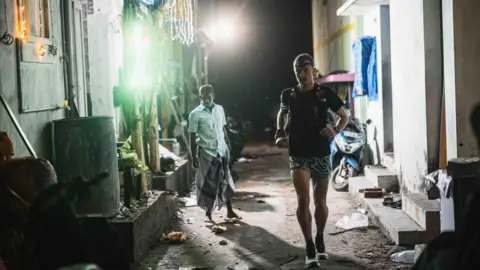 James Poole Will Robinson running in the dark through a back alleyway. He is wearing patterned shorts, a black t-shirt, a running vest and black backwards cap. The alley is dirty and littered, with a bright light on the left. A local Indian man wearing a blue buttoned shirt and wrap on his lower half is watching Will run past. 