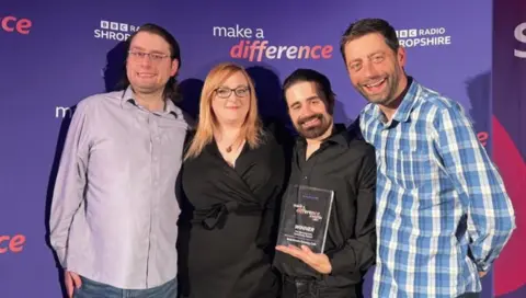 A man with short dark hair, glasses in a pale purple shirt, next to a woman with long blonde hair and a black dress, a man with dark hair and a beard in a black shirt and a man with short dark hair and a blue chequed shirt in front of a purple background