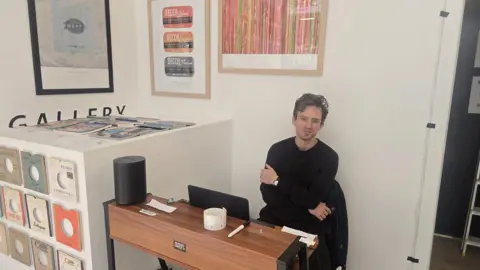 A person sits at a small wooden desk in a minimalist, gallery-like room with white walls. The person wears a dark long-sleeve top and sits with arms crossed, facing the camera. On the desk are a laptop, a white mug with a small pattern, and a pen. To the left, a display cabinet holds rows of vinyl records with colourful labels, and a stack of magazines or records is arranged on top. Several framed artworks hang on the wall behind, including designs featuring record labels and abstract patterns.