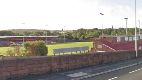 Google Borough Park from a nearby road. It's got two covered stands on either side of a large, green pitch. There are six tall floodlights and lots of trees behind the stadium in the distance.