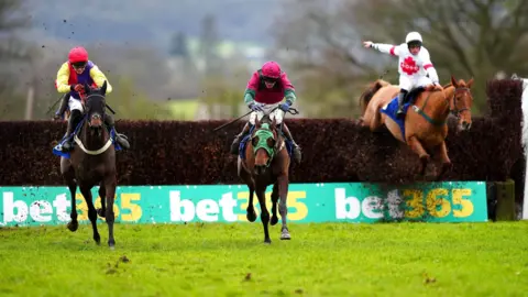 Three horses with their riders racing over a hurdle at Taunton race course. The jockeys are wearing bright yellow and pink tops.