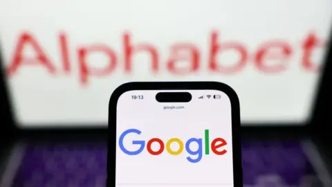 A mobile phone with the multi-coloured google logo on it, and a laptop with the red Alphabet logo in the background.