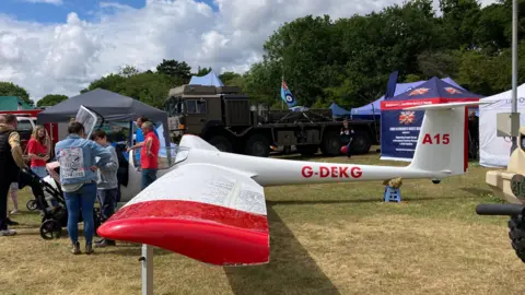 Luke Deal/BBC A glider sits in a grass field at the Suffolk Show. It is white with red tipped wings and its aircraft number painted in red along its body. People crown around its cockpit to take a look inside.