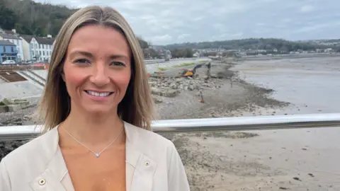 Local councillor Francesca O’Brien smiling at the camera near the Mumbles seafront, where a large digging machine is working on the beach