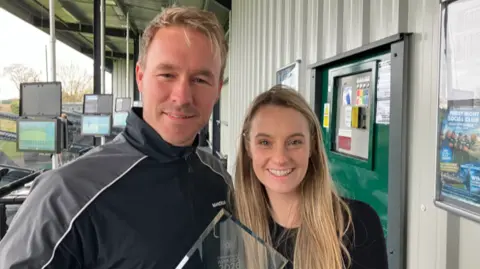 A man and woman, both with blonde hair, smile at the camera. They are holding a glass award between them. Behind them are bays of a golf driving range