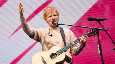 Ed Sheeran, in a beige T-shirt holding a guitar on a stage in front of a pink and white patterned background