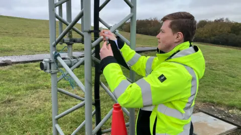 SAM READ/BBC A side on shot of Robert Forster who is wearing a hi-vis coat. He has his arms in the air holding a small plastic bottle which he has taken from a clip on a metal tower. He is also holding a clipboard. Behind him is a grass bank and there are some bushes in the background. There is a cloudy overcast sky overhead.
