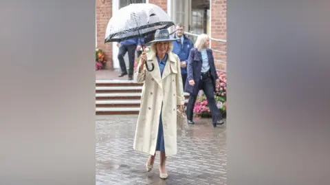 PA A person walks across a wet brick-paved courtyard holding a large transparent umbrella during rainfall. The person is wearing a light-coloured trench coat over a blue outfit, a wide-brimmed hat with decorative trim, and light-coloured shoes, while carrying a small handbag. Behind the person, steps lead up to a brick building with white-framed doors and windows. Several other people in coats are walking nearby, and bright flower displays are visible along the edges of the steps. Raindrops can be seen falling throughout the scene, and the reflective pavement emphasizes the rainy weather.