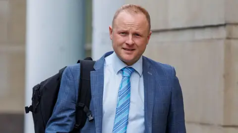 Jamie Bryson, a man with short, blonde hair, pictured outside court at an earlier hearing in the case.  He is wearing a light blue checked suit, a pale shirt and a striped blue tie.  He is carrying a black backpack on one shoulder. 
