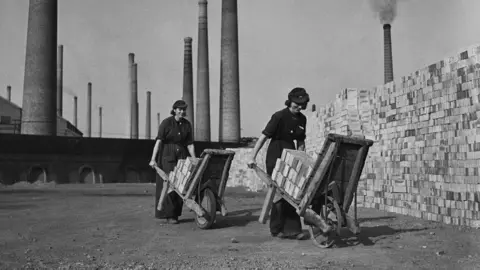 Two workers are pushing wheelbarrows loaded with bricks at the Stewartby brickworks. Large stacks of neatly arranged bricks line the right side of the scene.