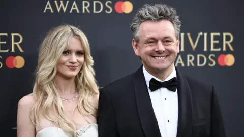 Getty Images Michael Sheen and wife Anna in formal dress, posing and smiling in front of an Olivier Awards-branded backdrop