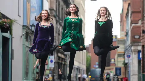 Belfast City Council Three women in traditional Irish dancing dresses - jewel toned velvet with celtic embellishments. They are pictured dancing, mid leap in the air on a city street. 