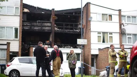 fire fighters and other people stand outside a building which has been severely damaged by a fire