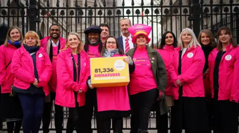 PA Media A group of 13 people stand by the gates for Downing Street wearing pink jackets and Theo Burrell is in the middle holding a yellow box that reads "81,336 signatures demanding change"