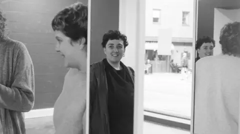 Haley Drolet Zahra is reflected in some tall mirrors. She has short dark hair and wears a dark shirt and jacket. Two other people are also partially reflected, possibly in conversation with her. The street outside can be glimpsed in the background, with people doing their shopping.