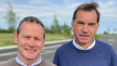 John Quinn (left) is standing at the cemetery. He has short brown hair and is wearing a blue shirt and beige jumper. Gerry Quinn is standing beside John and is wearing a blue jumper and white shirt.