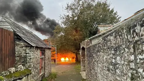 A fire blazes behind a large tree, down a small lane with high granite walls. Smoke billows in the background.