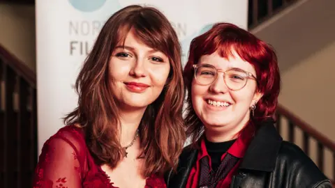 Khandie Photography Two people, smiling at the camera, at a film festival. The woman on the right has long dark hair and has a red top on and necklace, the person to the left has red shoulder-length hair, glasses on, a black leather jacket, red top and earrings in their ears. 