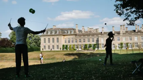 Tom Bradely/Greenbelt Boughton House stately home set behind a large dipped lawn. Two children in the foreground play with circus toys. There are no big crowds in this photo. The sun is shining.