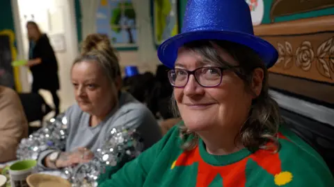 Two women are seated at a table during a festive gathering. Carrie in the foreground, wears a bright blue glittery top hat and a green jumper with a red and yellow Christmas design. The table is set with cups, plates, and silver tinsel decorations, and a colorful mural is visible in the background. The second woman has blonde hair in a bun and is wearing a grey and silver sparkly top.
