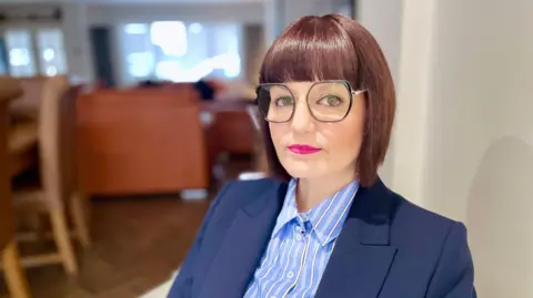 Mark Norman/BBC A woman with glasses and short dark hair, wearing a blue jacket and striped blue blouse.