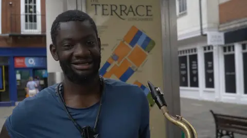 Meghan Shaw/LDRS A man in a blue shirt and holding a saxophone. He is standing at a high street.