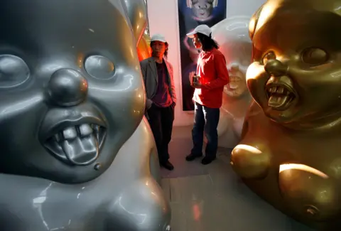 Reuters The Gao Brothers, both wearing white caps, stand between three of their "Miss Mao" sculptures. The sculptures are silver, gold and white and show a caricature-like Mao Zedong with buck-teeth, breasts and their tongues sticking out.