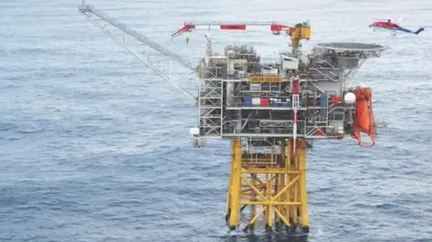Harbour Energy Oil and gas platform with yellow legs in the sea, a blue, red and white offshore helicopter hovers overhead, and vessels in the background.