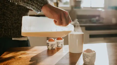 Getty Images A man's hand pouring milk into a steaming mug with boiled eggs on the wooden worktop