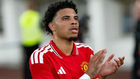 Ethan Williams in a red Manchester United shirt. He is clapping his hands to address fans in the crowd