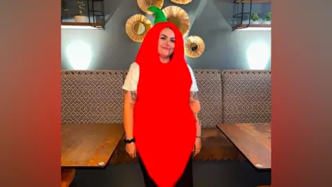 A woman smiling with a white t-shirt on and tattoo sleeve. She is dressed in a large red chilli costume which covers her head and hair and torso