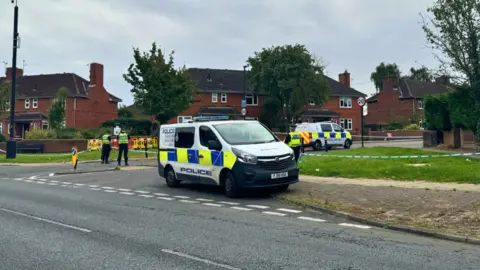 BBC/Emily Johnson Police officers and a van stand in front of a blue and white barrier at the top of Bellfarm Avenue 