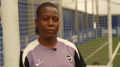 A woman wearing a purple, white and blue Brighton & Hove Albion training shirt is standing in goal at a training ground.